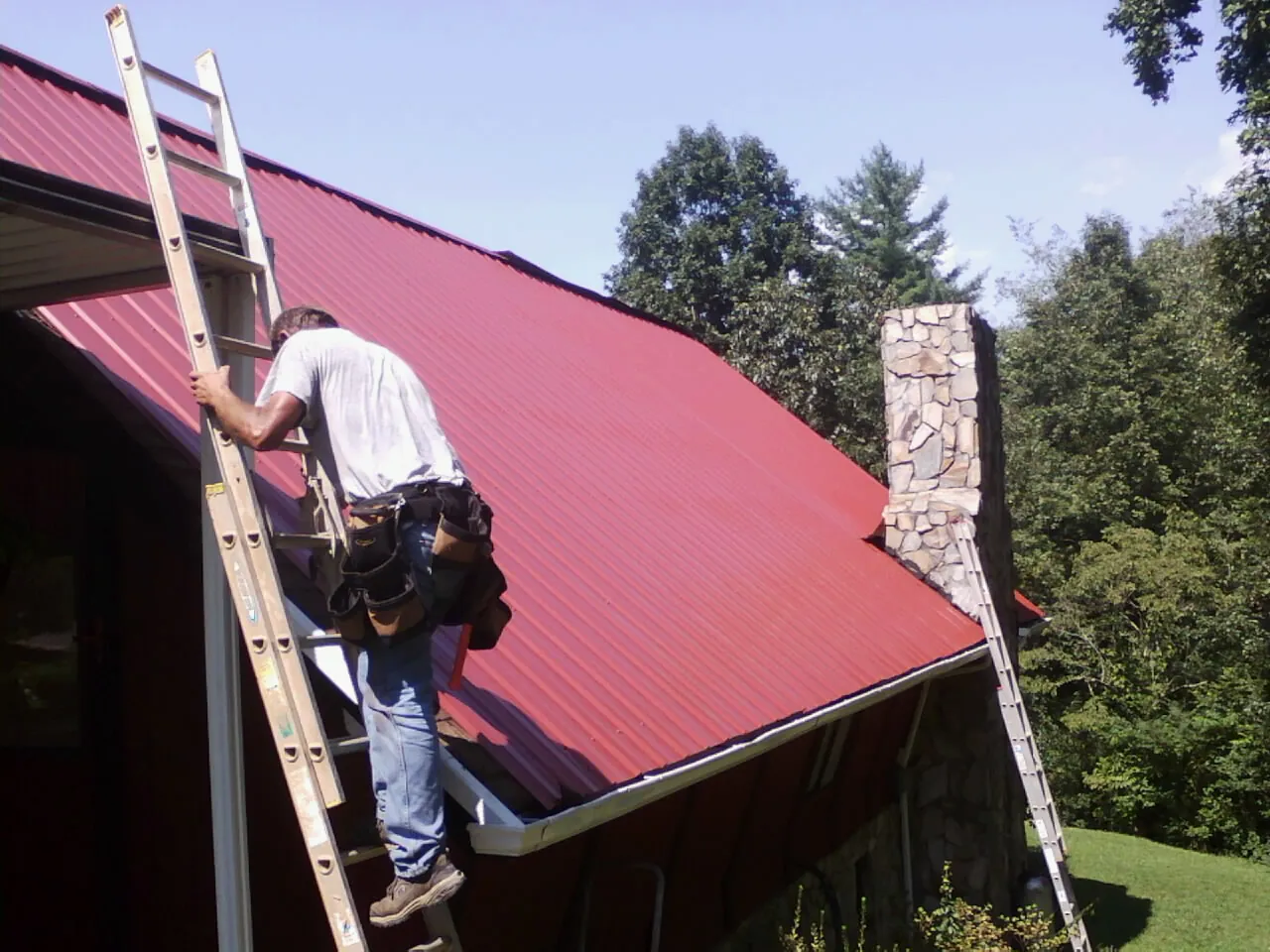 Roofer on a ladder beside a red metal roof and stone chimney — Soffit & Fascia Repair by Roofing Brothers in Bealeton